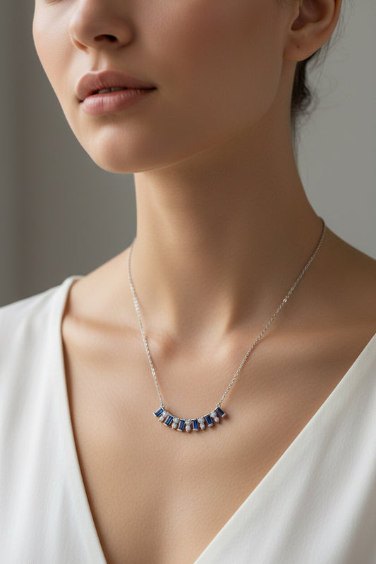 A silver necklace featuring a 1 inch curved bar pendant with multiple rectangular blue 3/8 inch baguette-cut stones and small clear accent stones, on chest
