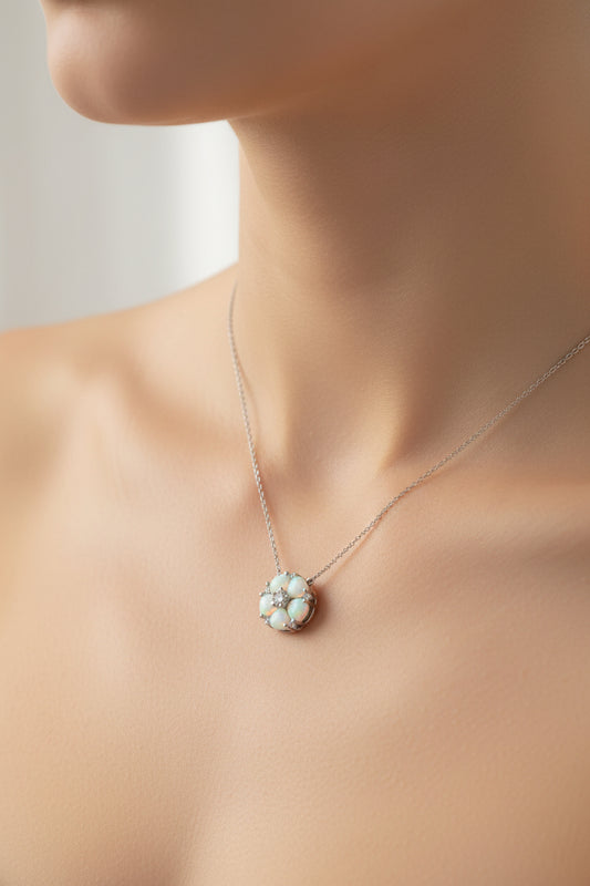 A close-up of a woman's neck and upper chest wearing a delicate silver chain necklace with a 1/2 inch in diameter flower-shaped pendant made of iridescent white opal-like stones and a small clear center stone.