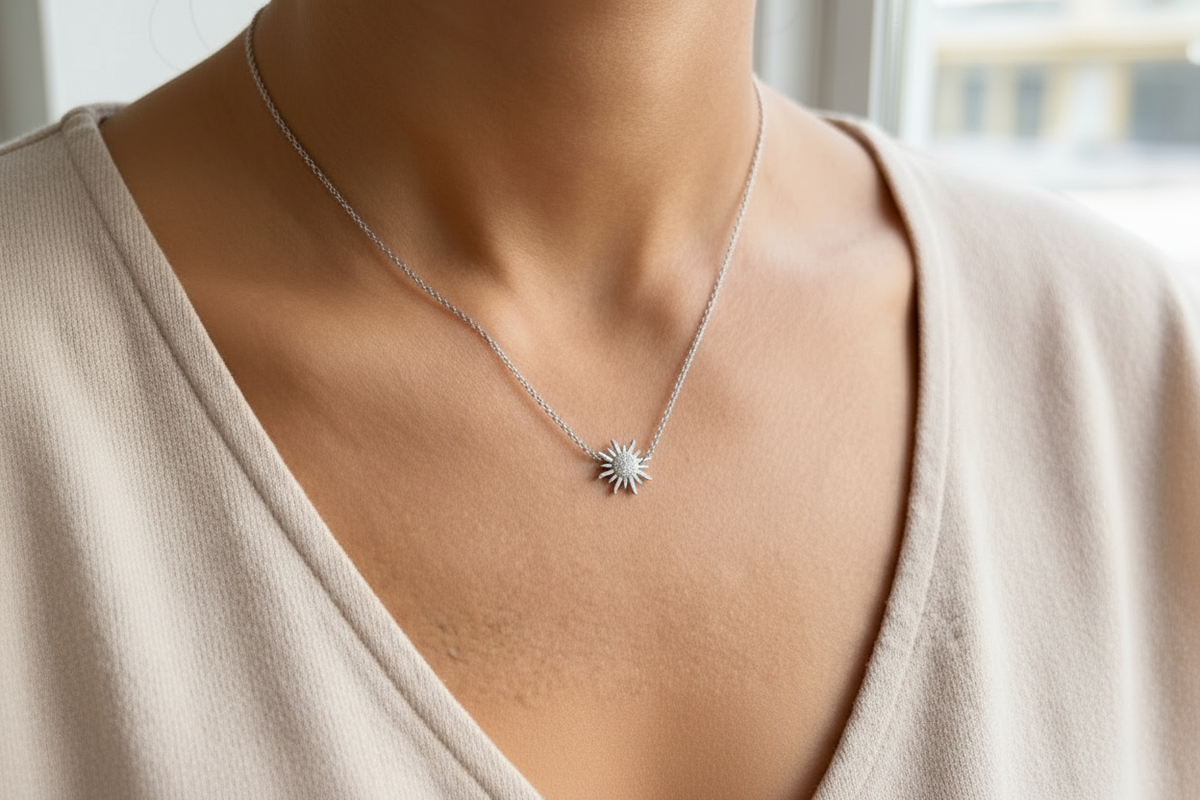 A close-up of a person's neck and chest wearing a delicate silver chain necklace with a sunburst pendant. The pendant features a textured center surrounded by radiating points, photographed in natural light. Very small