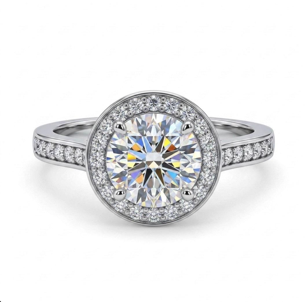 A round halo engagement ring featuring a large brilliant-cut center stone surrounded by a double halo of smaller stones, set in a silver-colored metal band with pavé accents on the shoulders, shown on a white background.